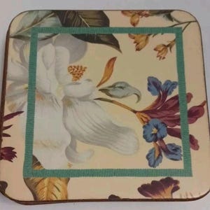 May include: Square coaster with a floral design. The coaster features a large white flower with yellow stamens, blue and purple flowers, and green leaves against a cream background. A teal border frames the floral design.