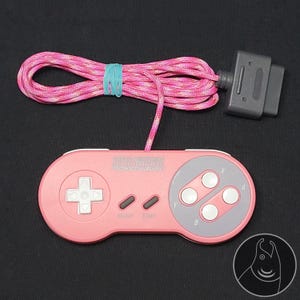 Könnte beinhalten: Ein rosafarbener Videospiel-Controller mit einem weißen Steuerkreuz und vier weißen Aktionstasten, die mit A, B, X und Y beschriftet sind. Der Controller hat ein graues Mittelteil, ein rosa-weiß geflochtenes Kabel und einen grauen Stecker.