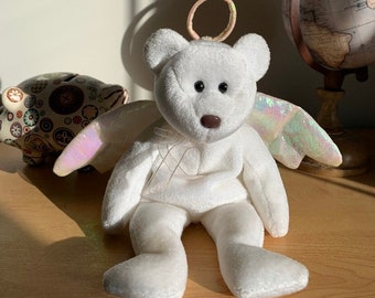 May include: White plush teddy bear with iridescent wings and a halo. The bear is sitting on a wooden surface.