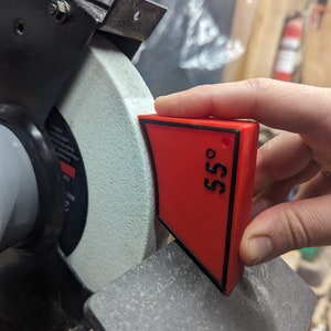 May include: A red plastic angle guide with a black border and the number "55°" printed on it. The angle guide is being held against a white grinding wheel on a bench grinder.