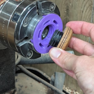 May include: A purple plastic ring with a black rubber insert is attached to a metal lathe chuck. A wooden knob is being held in the hand, ready to be attached to the chuck.