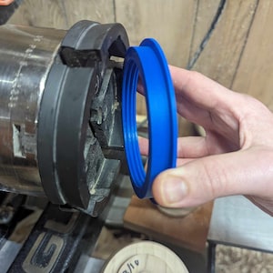 May include: A blue plastic ring with multiple grooves is being held up to a black metal lathe chuck. The ring is being used to create a wooden bowl.