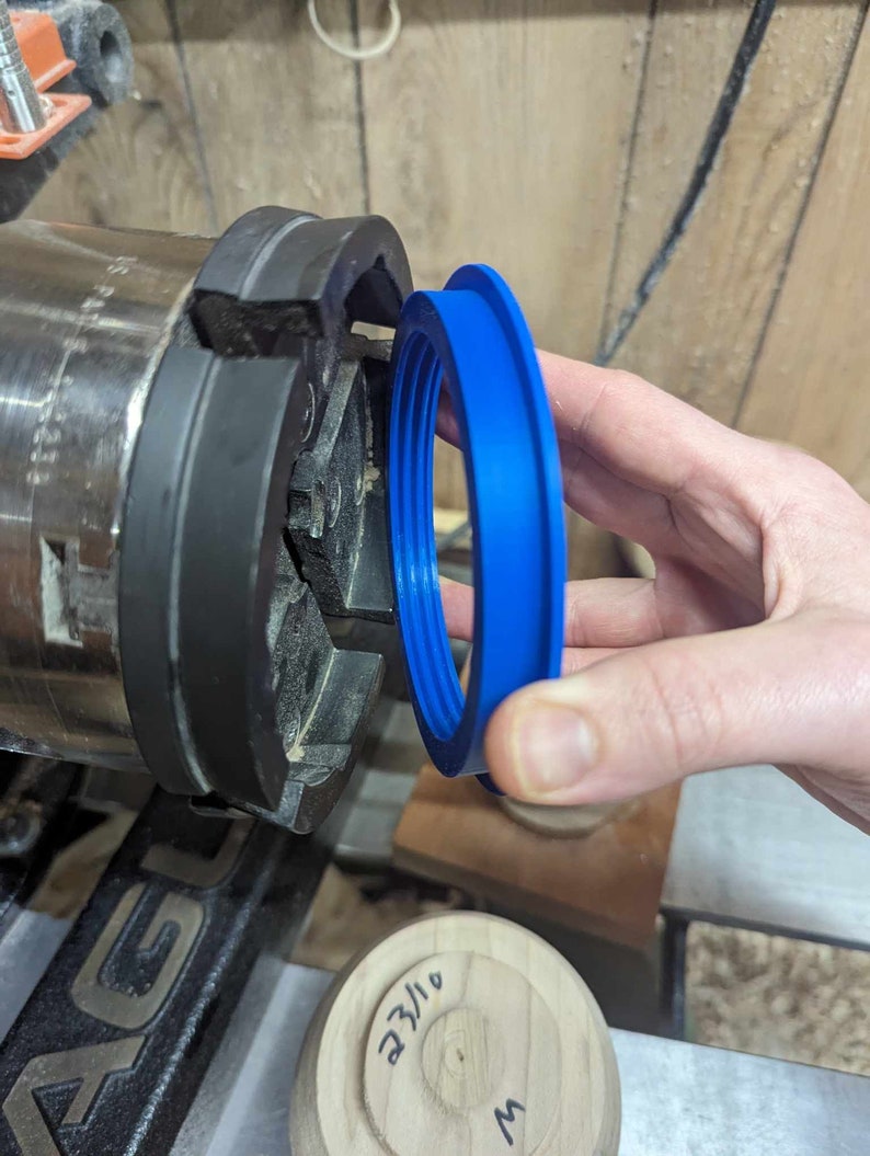 May include: A blue plastic ring with multiple grooves is being held up to a black metal lathe chuck. The ring is being used to create a wooden bowl.