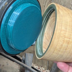 May include: A wooden bowl with a lid being turned on a lathe. The bowl is light brown and the lid is dark green.