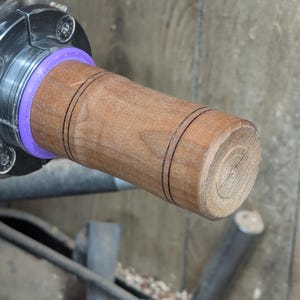 May include: A close-up of a wooden handle with two black lines around the center. The handle is being turned on a lathe.