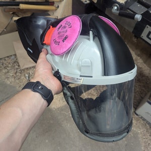 May include: A white and black safety helmet with a clear face shield and two pink 3M 2097 filters attached. The helmet has a black strap and a white label that says "trer".