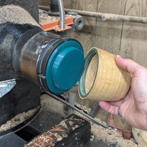 May include: A wooden bowl being turned on a lathe. The bowl is light brown and has a teal colored ring around the top. The lathe is black and has a teal colored chuck.