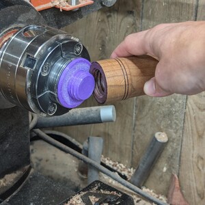 May include: A close-up of a wooden dowel being turned on a lathe. The dowel is being held in place by a purple plastic chuck. The dowel has a series of grooves carved into it.
