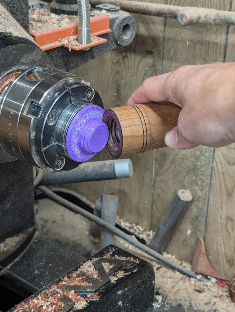 May include: A close-up of a wooden dowel being turned on a lathe. The dowel is being held in place by a purple plastic chuck. The dowel has a series of grooves carved into it.