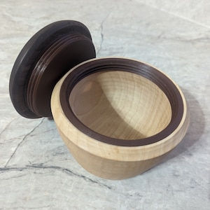 May include: A wooden bowl with a dark brown lid. The bowl is made of light wood and has a dark brown rim. The lid is made of dark wood and has a smooth surface.