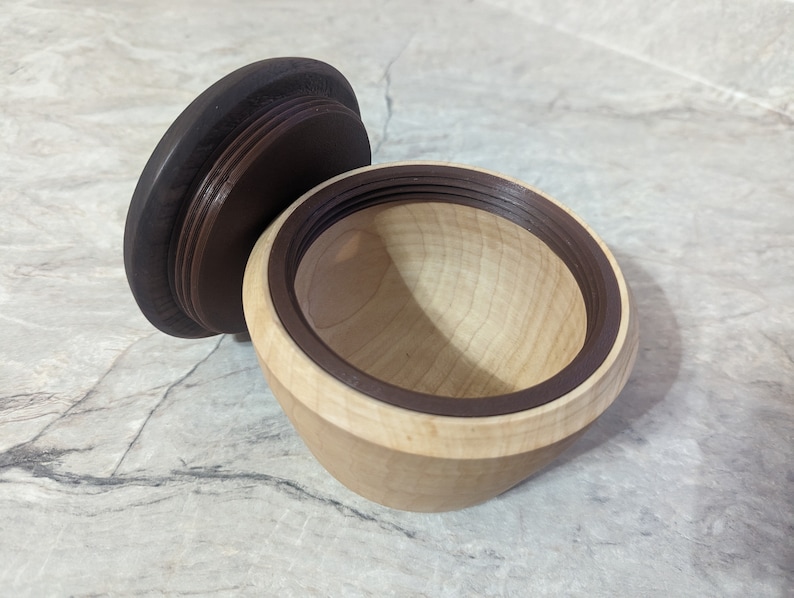 May include: A wooden bowl with a dark brown lid. The bowl is made of light wood and has a dark brown rim. The lid is made of dark wood and has a smooth surface.