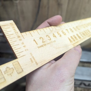 Op de afbeelding: Een houten liniaal met een esdoornbladlogo, gemarkeerd in inches en een halve schaal diametermeting. De liniaal is gelabeld "ASH & EWAER" en "DIAMETER (HALF SCALE)".