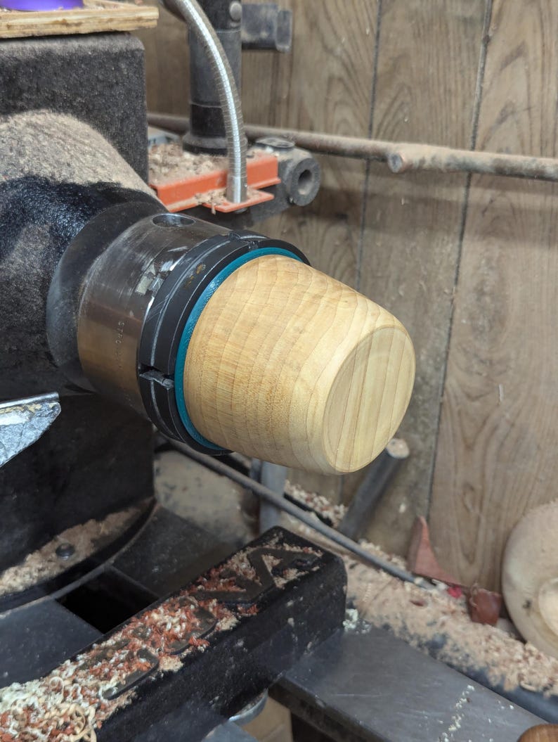 May include: A close-up of a wooden bowl being turned on a lathe. The bowl is light brown and has a smooth finish. The lathe is black and has a metal chuck.
