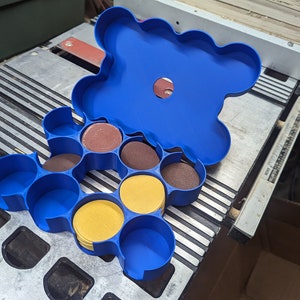 May include: A blue plastic storage container with multiple compartments for holding round sanding discs. The container is open and the compartments are filled with yellow and brown sanding discs.