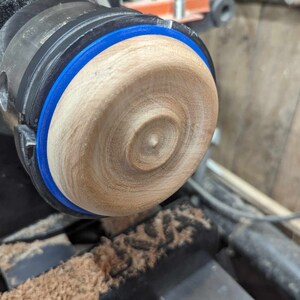 May include: A close-up of a wooden bowl being turned on a lathe. The bowl is being shaped with a blue tape measure around the edge.
