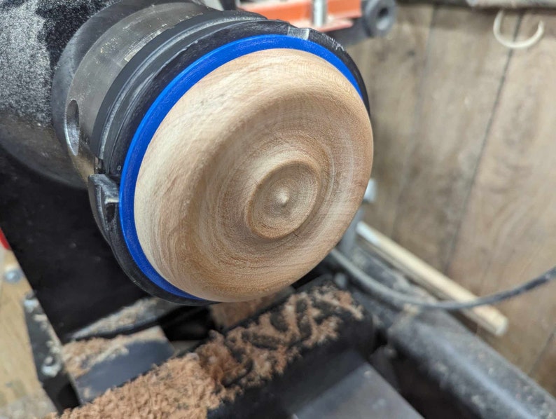 May include: A close-up of a wooden bowl being turned on a lathe. The bowl is being shaped with a blue tape measure around the edge.
