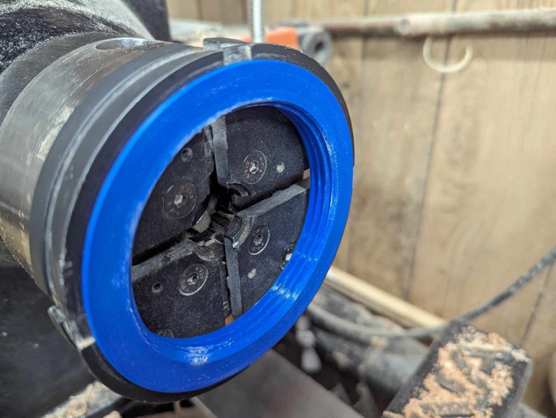 May include: Close-up of a blue and black lathe chuck with four jaws. The jaws are open and ready to hold a piece of wood.