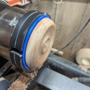 May include: A close-up of a wood turning project on a lathe. The wood is light brown and has a blue ring around the edge. The lathe is black and silver.