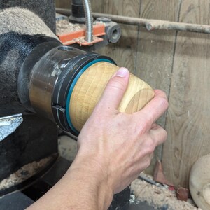May include: A close-up of a wooden bowl being turned on a lathe. The bowl is being held in place by a chuck and is being turned by a hand.