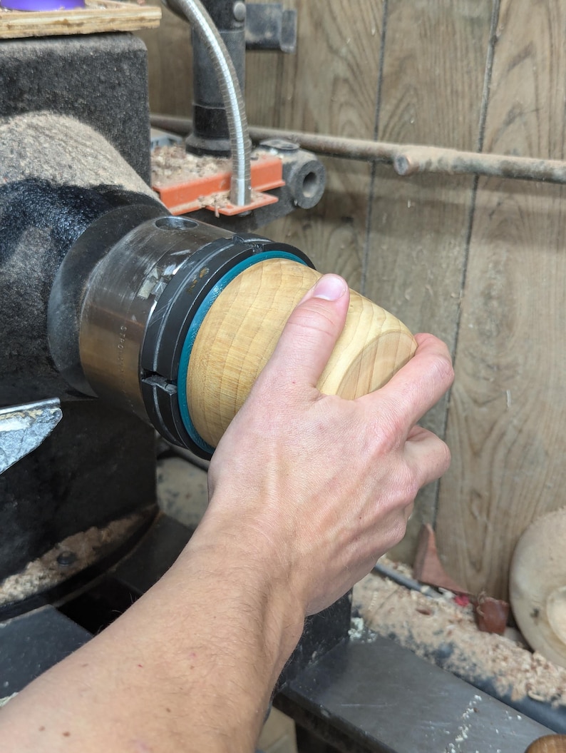 May include: A close-up of a wooden bowl being turned on a lathe. The bowl is being held in place by a chuck and is being turned by a hand.