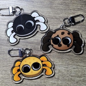 May include: Three different spider-shaped keychains with googly eyes. The first keychain is black with white legs, the second is brown with brown legs, and the third is yellow with yellow legs.
