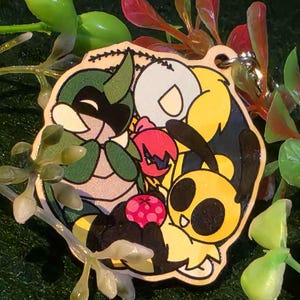May include: A wooden keychain featuring a colorful cartoon design. The design includes a green insect-like character, a yellow and black bee-like character, and a red fruit. The keychain has a silver-colored metal ring.