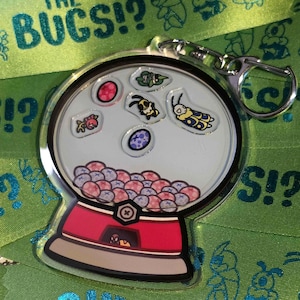 May include: A clear acrylic keychain shaped like a gumball machine. The keychain features a red base, a clear globe with various bug illustrations, and the text "THE BUCS!?" on a green ribbon.