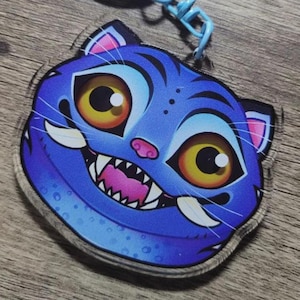 以下が含まれることがあります： 青い猫の顔のキーホルダー。漫画のデザインで、大きな黄色の目、ピンクの鼻、歯を見せた笑顔が特徴です。キーホルダーは水色のチェーンに付いています。