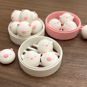 May include: A set of three white ceramic pig-shaped steamers with pink details. The steamers are in different sizes and are arranged in a row on a wooden surface.