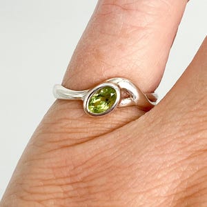 Può includere: Un anello in argento con una singola piccola pietra preziosa di peridoto di colore verde chiaro, di forma ovale e incastonata in una lunetta liscia. La fascia ha una finitura metallica lucida e riflettente con un design curvo.