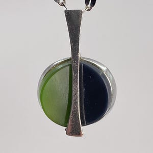 Op de afbeelding: Een zilverkleurige hangerketting met een ronde bedel verdeeld in twee helften, één limoengroen en één donker marineblauw, gescheiden door een verticale zilverkleurige staaf. De hanger hangt aan een fijne zilverkleurige ketting met ovale schakels.
