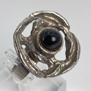 Op de afbeelding: Een zilverkleurige ring met een cirkelvormig, getextureerd, opengewerkt frame met een gladde, gepolijste, ronde zwarte steen in het midden. Het metalen oppervlak heeft een ruwe, onregelmatige afwerking en de ringband is eenvoudig en strak.