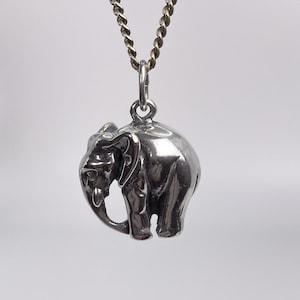 Puede incluir: Un colgante de elefante en tono plateado pulido suspendido de una cadena de metal trenzado a juego. El dije de elefante presenta un diseño tridimensional con orejas detalladas, una trompa curva y un acabado de superficie liso y reflectante.