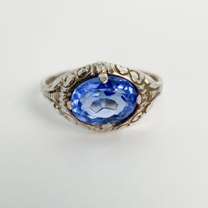 Op de afbeelding: Een zilverkleurige ring met een centrale ovaal geslepen blauwe edelsteen. De metalen band is versierd met ingewikkelde gegraveerde krulpatronen rondom de steen, die op zijn plaats wordt gehouden door kleine pootjes aan de boven- en onderrand.