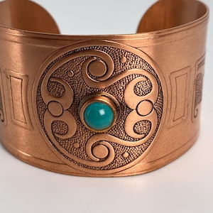Op de afbeelding: Een brede koperen manchetarmband met een centraal rond medaillon met reliëf van wervelende patronen en een getextureerde achtergrond. Een gladde, ronde, helder turquoise steen is in het midden van het medaillon gezet. Gepolijste afwerking.