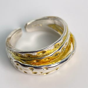Könnte beinhalten: Ein offener Ring mit mehrschichtigem Design, bestehend aus einem polierten silberfarbenen Außenring und einem texturierten, vertieften goldfarbenen Innenring. Die Silberoberfläche weist kleine kreisförmige Vertiefungen auf.