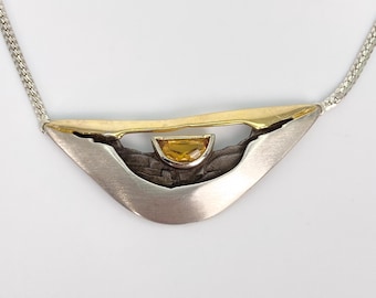 May include: A necklace featuring a crescent-shaped pendant made of brushed silver and gold-tone metal. The center of the pendant contains a small, semi-circular yellow gemstone set within a dark, textured, oxidized silver background.