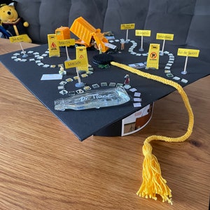 Peut inclure: Un bonnet de graduation noir avec un gland jaune. Le bonnet est décoré d'un chemin de ficelle jaune, de panneaux de chantier miniatures et de figurines de jouets. Les panneaux portent les noms de villes : Jundiaí, Brasília, Wittlich, Campes, Sab Sestion, Braunting et Rio de Janeiro. Le bonnet a également une petite plaque de verre avec le texte "Dr. Hane".