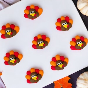 May include: A collection of nine colorful turkey-shaped decorations. Each turkey has a brown body, red and orange tail feathers, and a yellow beak. The decorations are arranged on a white surface, with fall-themed accents.