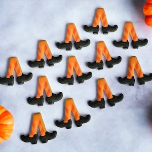 May include: A collection of orange and black witch leg decorations with black shoes and orange and white striped stockings. Two small orange pumpkins are also in the image. The decorations are likely for Halloween.
