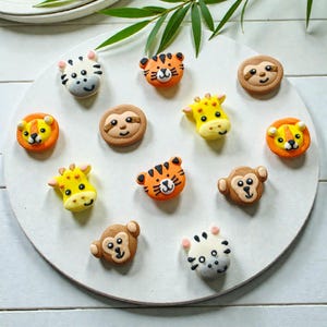May include: A collection of colorful, animal-shaped cookies, including a zebra, tiger, sloth, giraffe, lion, and monkey. The cookies are arranged on a white, circular plate, with a sprig of greenery in the background. The cookies are likely for a party or celebration.
