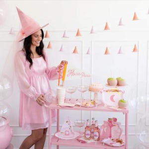 May include: A pink witch costume with a witch hat and veil, a pink and white dessert table with a sign that says "Hocus Pocus", and pink and white balloons.