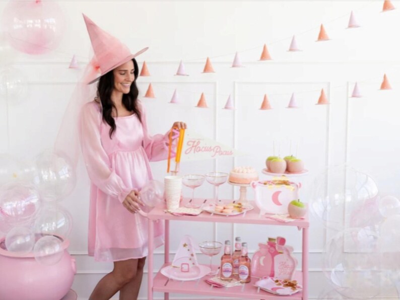 May include: A pink witch costume with a witch hat and veil, a pink and white dessert table with a sign that says "Hocus Pocus", and pink and white balloons.