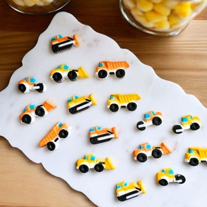 May include: A collection of colorful, miniature construction vehicles, including bulldozers, dump trucks, and front-end loaders. The vehicles are orange, yellow, and blue, arranged on a white, marble-like surface. The image has a playful, whimsical feel.