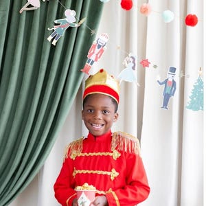 May include: A child dressed in a red and gold Nutcracker costume, holding a popcorn container with a Nutcracker illustration. The background includes a garland of Nutcracker-themed cutouts and pom-poms, with green and white curtains.