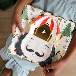 May include: A Nutcracker themed paper plate with a gold and red hat, black hair, a white face, and a black mustache. The plate has a white background with a green Christmas tree and other festive designs.