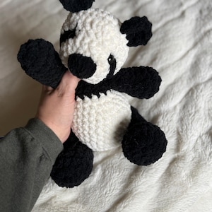 May include: A handmade crochet panda bear with black and white yarn. The panda has black ears, arms, and legs, with a white body and head. The panda's face has black eyes and a small smile. This soft toy is perfect for a child's room.