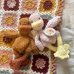 May include: Two handmade crochet duck toys in shades of brown and pink, with yellow beaks and accents, resting on a colorful granny square blanket. The blanket features squares of various colors, including red, pink, and yellow, with white borders.
