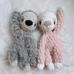 May include: Two handmade crochet sloths. One sloth is gray with a black nose and eyes, the other is pink with a tan nose and black eyes. Both sloths have white paws and are laying on a white, fluffy surface.