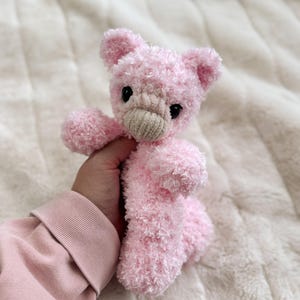 May include: A handmade pink teddy bear with black eyes and a beige muzzle. The plush toy is made of soft, fluffy material and has small ears and stubby arms and legs. The bear is held against a white, textured background.